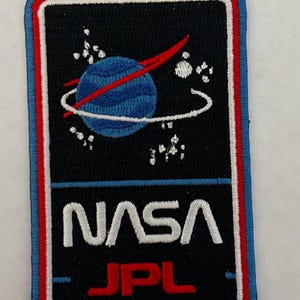 Könnte beinhalten: Ein schwarzer gestickter Aufnäher mit einem blauen und roten NASA JPL-Logo. Das Logo zeigt einen blauen Planeten mit einer roten Umlaufbahn darum, umgeben von weißen Sternen.