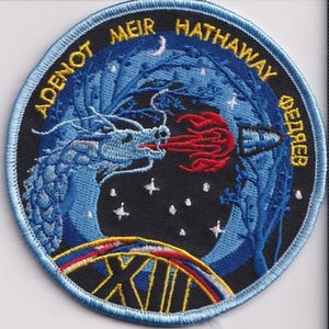 May include: Embroidered patch with a blue dragon breathing red flames at a rocket. The patch has a black background with stars and a crescent moon. Text around the edge reads "ADENOT MEIR HATHAWAY ФЕДЯЕВ" and "XIII".