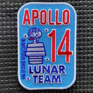 May include: A blue embroidered patch with the text "APOLLO 14 LUNAR TEAM" and a cartoon image of Snoopy in a space suit standing on a lunar module.