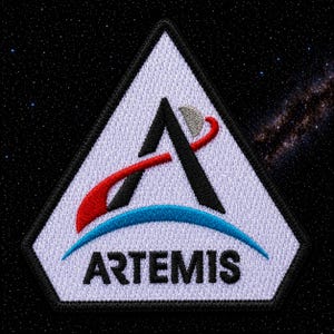 Peut inclure: Écusson brodé avec le logo du programme Artemis. Le design comprend un triangle blanc avec un 'A' noir et un trait rouge et bleu. Le mot 'ARTEMIS' est en noir, en dessous du logo, sur fond étoilé.