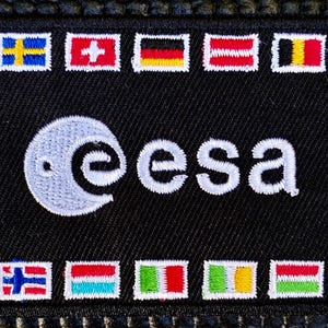 Pode incluir: Patch bordado preto com o logotipo branco da ESA e bandeiras de diferentes países. Este patch retangular é provavelmente uma lembrança ou emblema, possivelmente para uma agência espacial.