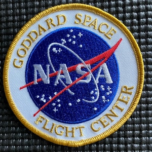 May include: A white and gold embroidered patch with the NASA logo and the text "Goddard Space Flight Center".