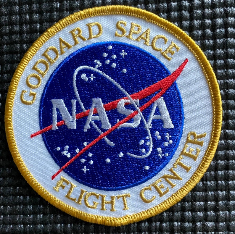 May include: A white and gold embroidered patch with the NASA logo and the text "Goddard Space Flight Center".
