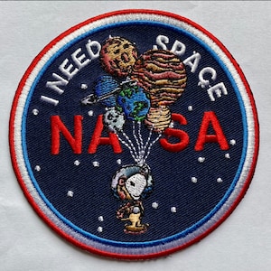 Puede incluir: Un parche bordado azul con un borde blanco y rojo. Presenta un personaje de dibujos animados sosteniendo un montón de globos coloridos que dicen "Necesito espacio" y "NASA".