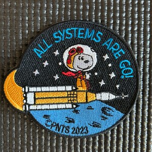 Puede incluir: Parche bordado con Snoopy con traje espacial en un cohete, con el texto "ALL SYSTEMS ARE GO!" sobre un fondo negro estrellado. El parche incluye el texto "©PNTS 2023".