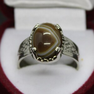 May include: A silver ring with a brown and white agate stone set in a claw setting. The ring has a textured band.
