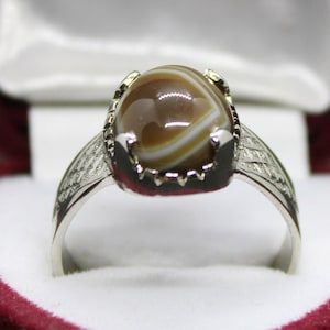 May include: A silver ring with a brown and white agate stone set in a silver bezel. The ring has a decorative band with a geometric pattern.