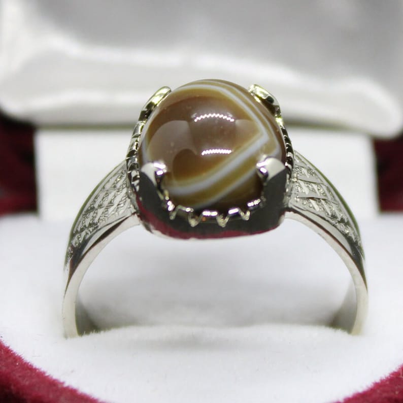 May include: A silver ring with a brown and white agate stone set in a silver bezel. The ring has a decorative band with a geometric pattern.