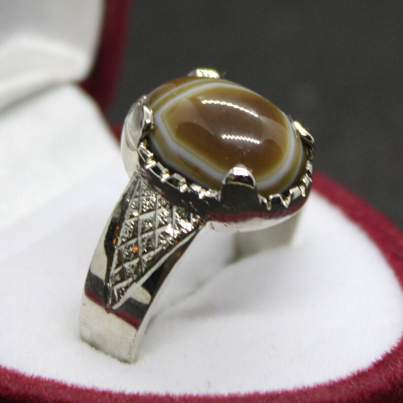 May include: A silver ring with a brown and white agate stone set in a decorative silver bezel. The ring band has a geometric pattern.
