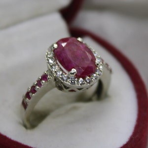 Pode incluir: Um anel de prata com uma grande pedra de rubi ovalada cercada por um halo de pedras brancas menores. A banda é adornada com rubis menores.