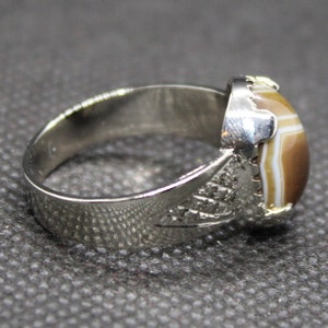 May include: A silver ring with a brown and white agate stone set in the center. The band of the ring is decorated with an intricate design.