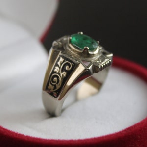 May include: A silver ring featuring a green emerald gemstone set in an ornate design. The band of the ring is engraved with a floral pattern.
