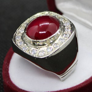 May include: A silver ring with a large, oval, deep red gemstone at the center, surrounded by a halo of smaller, clear stones. The ring has a wide band with a polished finish, set against a red velvet background.