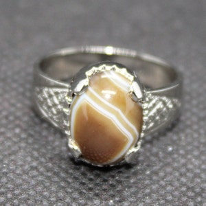 May include: A silver ring with a brown and white agate stone set in a silver bezel. The ring has a decorative design on the band.