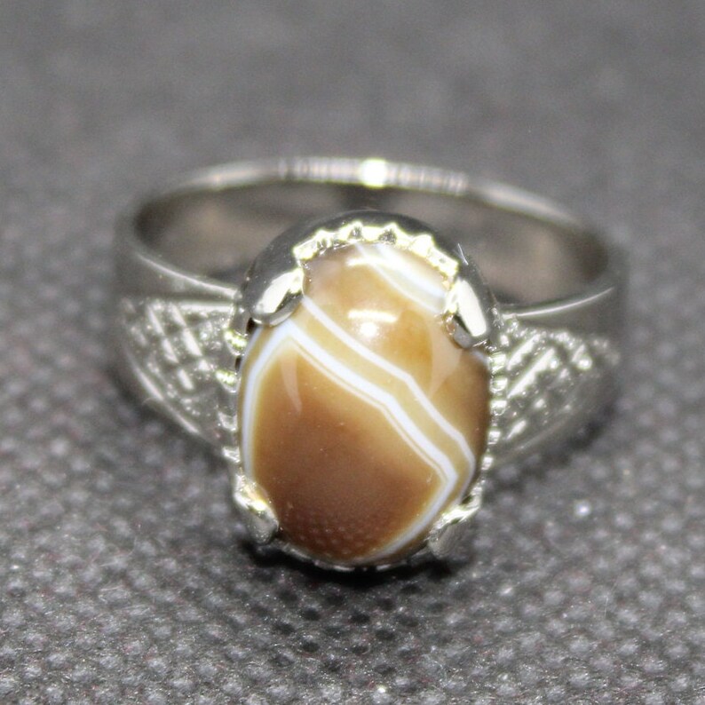 May include: A silver ring with a brown and white agate stone set in a silver bezel. The ring has a decorative design on the band.