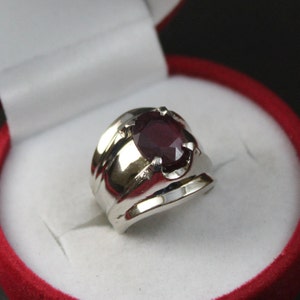 May include: A silver ring with a large, round, dark red gemstone set in a prong setting. The ring is in a red velvet ring box.