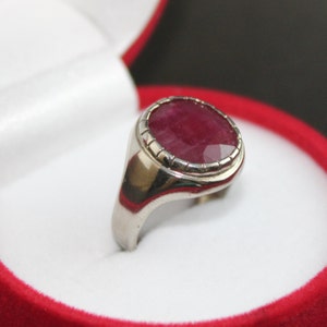 May include: A silver ring with a large, oval-shaped ruby gemstone set in a bezel setting. The ruby is a deep red color and has a faceted cut.