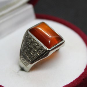 May include: A silver ring with a rectangular orange gemstone set in the centre. The band of the ring has a decorative pattern.