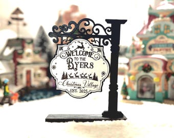 Può includere: Un cartello natalizio in bianco e nero con la scritta "Welcome to the Byers" e "Christmas Village EST. 2025". Il cartello presenta disegni di renne e fiocchi di neve. Il cartello è montato su un palo nero.