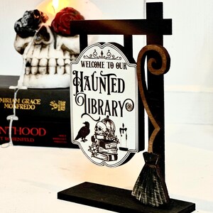May include: A wooden sign with a black and brown finish, shaped like a broom, with a white sign that reads "Welcome to our Haunted Library". The sign features a black raven, a skull, and a stack of books.