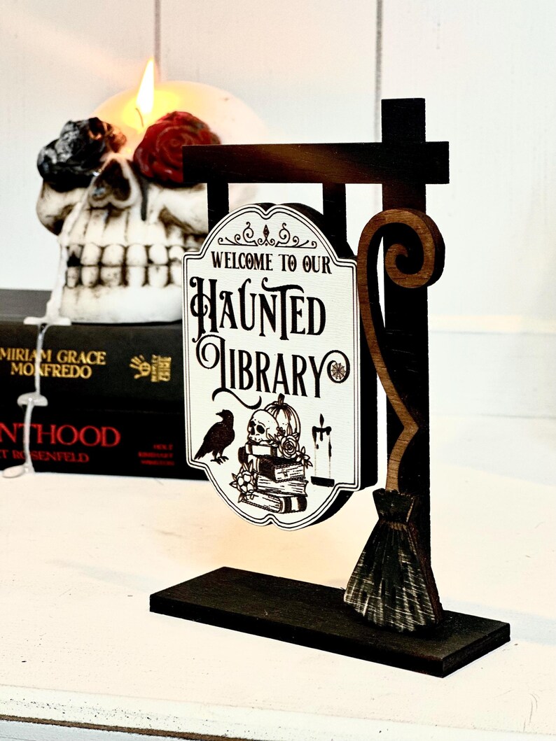 May include: A wooden sign with a black and brown finish, shaped like a broom, with a white sign that reads "Welcome to our Haunted Library". The sign features a black raven, a skull, and a stack of books.