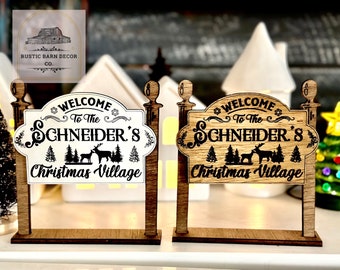 Può includere: Due cartelli in legno del villaggio di Natale. Un cartello è bianco con testo nero che recita "Benvenuti al villaggio di Natale di Schneider" con un cervo e una grafica ad albero. L'altro cartello è color legno con lo stesso testo e grafica.