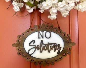 May include: Wooden oval sign with the words "NO Soliciting" in a decorative font. The sign is brown with a white center and is hanging on a door. White flowers are at the top of the image.