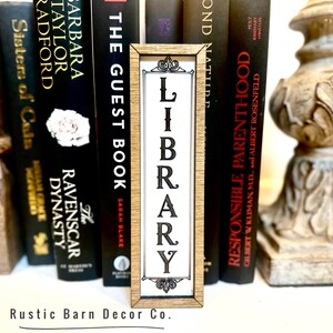May include: A wooden sign with the word "LIBRARY" in black letters on a white background. The sign is framed in a light brown wood frame and is sitting on a shelf with books.