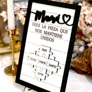May include: A black framed sign with a white background and the words "Mom, Eres la pieza que nos mantiene unidos" in black. The sign features a puzzle piece design with the names "Papa, Oliver, Mamac, Juniper, and Alma" written on each piece.