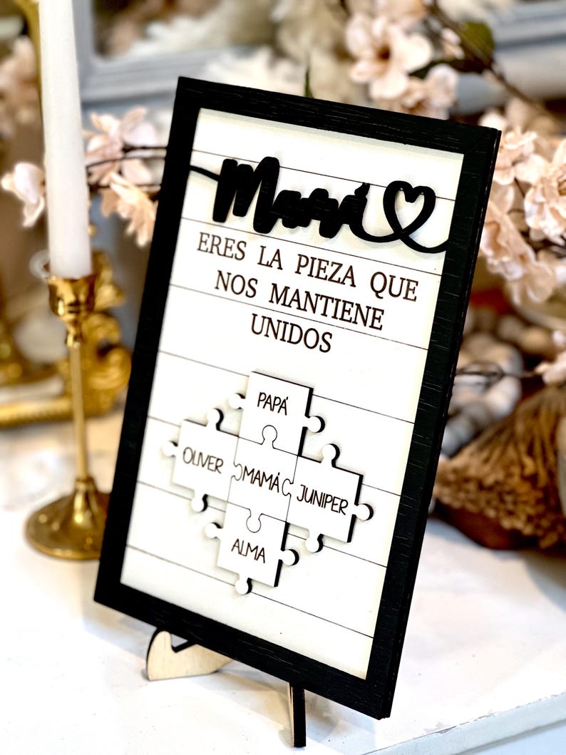 May include: A black framed sign with a white background and the words "Mom, Eres la pieza que nos mantiene unidos" in black. The sign features a puzzle piece design with the names "Papa, Oliver, Mamac, Juniper, and Alma" written on each piece.