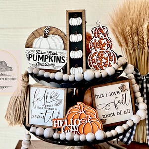 May include: A tiered tray decorated with fall-themed wooden signs, pumpkins, and wheat stalks. The signs read "Farm Fresh Pumpkins Open Daily", "Fall is proof that change is beautiful", and "But I love fall most of all."