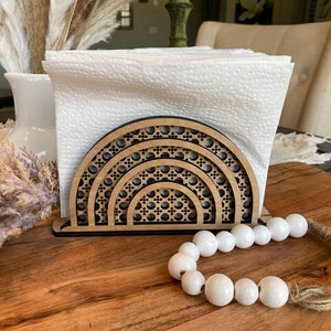 May include: A wooden napkin holder with a geometric design in the shape of a half circle. The holder is made of dark wood and has a black finish. It is holding a stack of white paper napkins. A string of white beads is in the foreground.