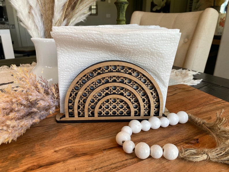 May include: A wooden napkin holder with a geometric design in the shape of a half circle. The holder is made of dark wood and has a black finish. It is holding a stack of white paper napkins. A string of white beads is in the foreground.