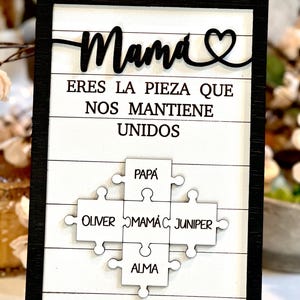 May include: A black framed sign with white wood paneling. The sign reads "Mama eres la pieza que nos mantiene unidos" with a heart symbol. Below the text are five white puzzle pieces with the names "Papa", "Oliver", "Mama", "Juniper", and "Alma" written on them.