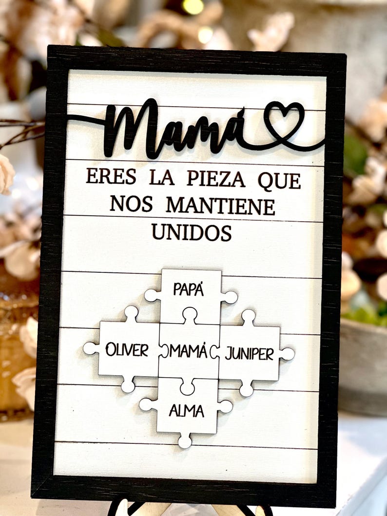 May include: A black framed sign with white wood paneling. The sign reads "Mama eres la pieza que nos mantiene unidos" with a heart symbol. Below the text are five white puzzle pieces with the names "Papa", "Oliver", "Mama", "Juniper", and "Alma" written on them.
