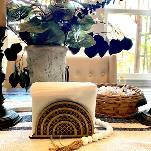 May include: A wooden napkin holder with a geometric design in a light brown color. The holder is on a white and black striped tablecloth with a white beaded garland and a brown tassel.