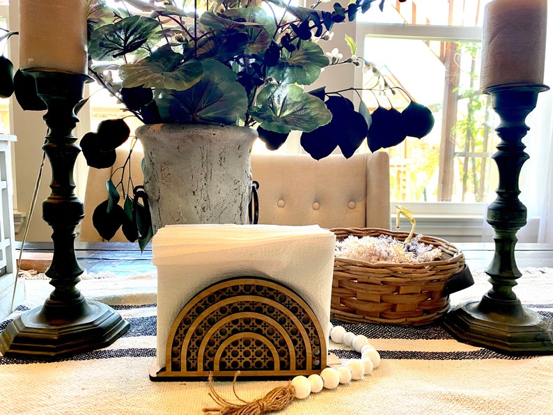 May include: A wooden napkin holder with a geometric design in a light brown color. The holder is on a white and black striped tablecloth with a white beaded garland and a brown tassel.