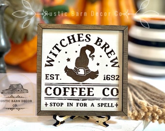 May include: A rustic wooden sign with a white background and black lettering. The sign reads "Witches Brew Est. 1692 Coffee Co. Stop in for a spell". A witch's hat with a buckle is depicted in the center of the sign.