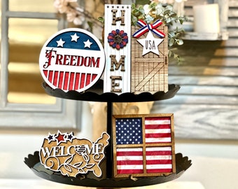 May include: A tiered tray decorated with patriotic themed wooden signs. The top tier features a round sign with the words "FREEDOM" and stars and stripes, a wooden sign with the word "HOME" and a star, and a wooden sign with the word "USA" and a barn door design. The bottom tier features a wooden sign with the words "WELCOME" and a map of the United States, and a framed picture of the American flag.