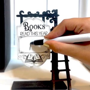 以下が含まれることがあります： 白地に「Books Read This Year」と書かれた白黒の看板。手は白いマーカーで看板に書いています。