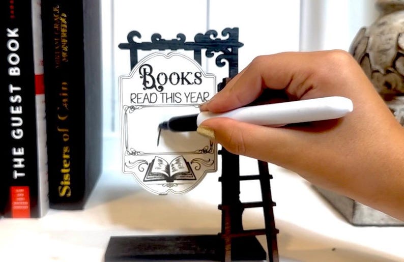以下が含まれることがあります： 白地に「Books Read This Year」と書かれた白黒の看板。手は白いマーカーで看板に書いています。