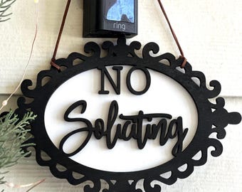 May include: A black wooden "No Soliciting" sign with an ornate oval frame hangs below a black doorbell camera. The sign has white lettering and is suspended by brown cords. The words "Rustic Barn Decor Co." are at the bottom.