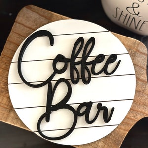 Pu&ograve; includere: Un cartello rotondo bianco con scritta in corsivo nero che recita "Coffee Bar" su una base in legno.