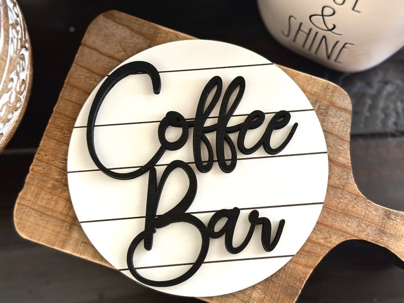 Pu&ograve; includere: Un cartello rotondo bianco con scritta in corsivo nero che recita "Coffee Bar" su una base in legno.