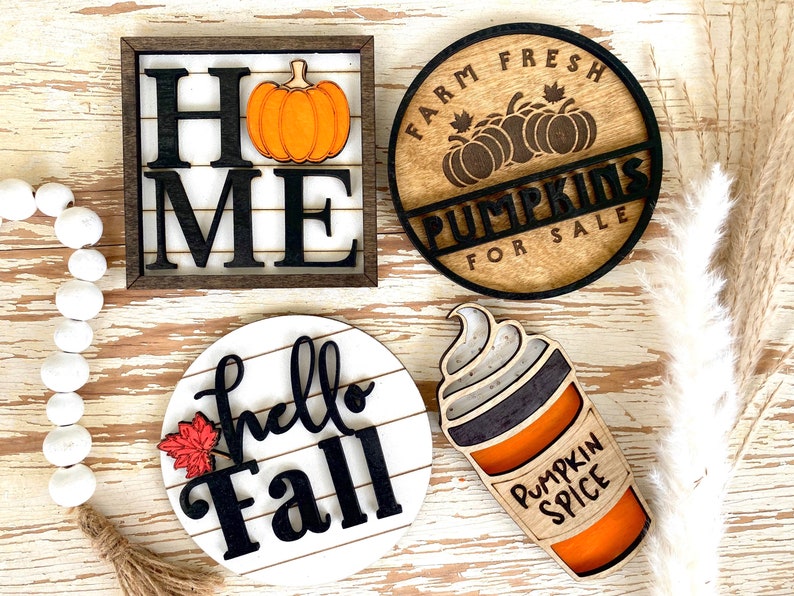 May include: Four wooden signs with fall themes. The first sign is a square sign with a brown frame and white background. The sign says 'Home' with a black pumpkin in the center. The second sign is a round sign with a brown frame and white background. The sign says 'Farm Fresh Pumpkins For Sale' with a black pumpkin in the center. The third sign is a round sign with a white frame and white background. The sign says 'hello Fall' with a red leaf in the corner. The fourth sign is a round sign with a brown frame and white background. The sign says 'Pumpkin Spice' with a brown and orange cup of coffee with whipped cream on top.