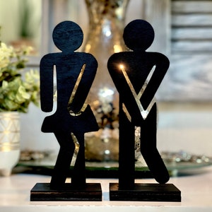 May include: Black wooden silhouette figures of a man and woman standing on separate black bases. The figures are stylized and represent the traditional restroom symbols.