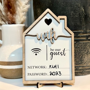 May include: Wooden sign shaped like a house with the word "wifi" in a cursive font. The sign also says "be our guest" and has a wifi symbol, network name "FAMILY", and password "2023".