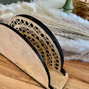 May include: A wooden napkin holder with a black and white geometric design. The holder is made of light wood and has a curved shape.
