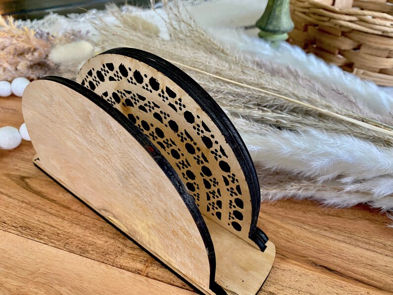 May include: A wooden napkin holder with a black and white geometric design. The holder is made of light wood and has a curved shape.