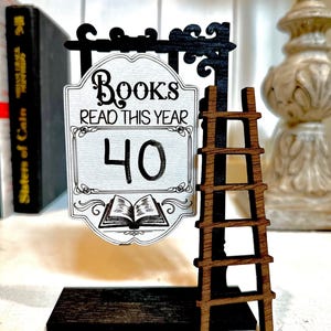 以下が含まれることがあります： 黒い梯子と「Books Read This Year 40」と書かれた白い看板が付いた木製看板。看板は黒い台座の上に置かれ、「Rustic Barn Decor Co.」の文字が書かれている